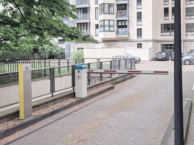 Eine Schrankenanlage mit automatischem Zugangssystem blockiert den Weg zu einem Parkplatz hinter einem Wohngebäude. Auf der linken Seite befindet sich ein Bedienfeld, während die Schranke geschlossen ist. Im Hintergrund sind mehrere parkende Autos und ein grüner Bereich mit Bäumen sichtbar.