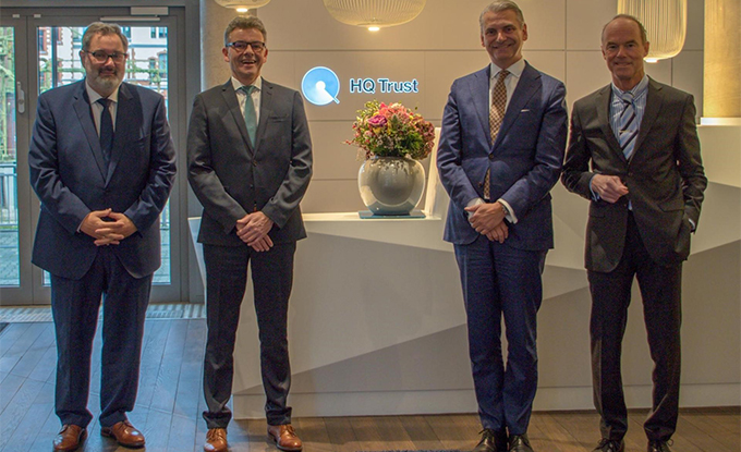 Vier Geschäftsleute in formeller Kleidung stehen nebeneinander vor einem Empfangstresen mit dem Logo von HQ Trust und einer Blumenvase.