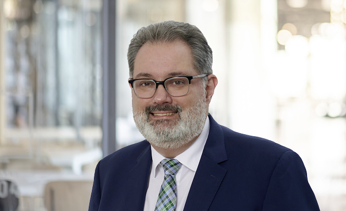 Ein Mann mit Brille und grauem Bart trägt einen dunklen Anzug und ein helles Hemd mit einer karierten Krawatte. Er lächelt freundlich vor einem unscharfen Hintergrund, der eine moderne Innenarchitektur zeigt.