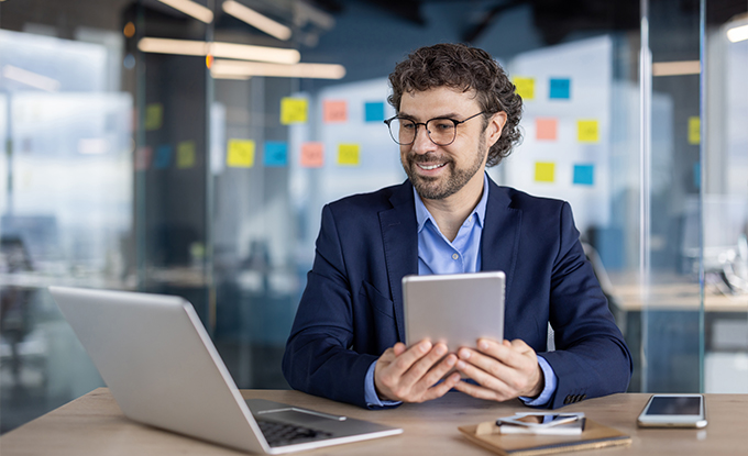 Ein lächelnder Mann mit Brille sitzt an einem Tisch und hält ein Tablet in der Hand, während er auf einen Laptop vor sich schaut. Im Hintergrund sind bunte Haftnotizen an einer Wand angebracht.