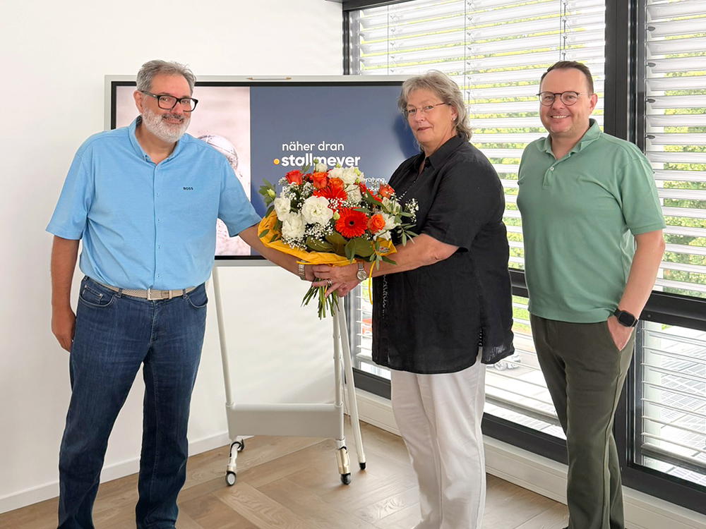 Eine Person überreicht einer anderen Person einen großen Blumenstrauß, während ein Dritter lächelnd daneben steht. Im Hintergrund ist ein Bildschirm mit dem Text „näher dran • stoll + never“ zu sehen.