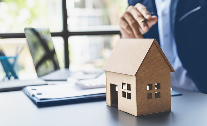 Ein Modelle eines Hauses aus Karton steht auf einem Tisch, während eine Person im Hintergrund mit einem Laptop arbeitet und auf das Modell zeigt.