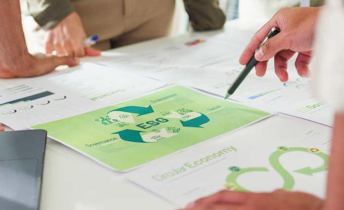 Auf einem Tisch liegen mehrere Dokumente, darunter eine Grafik mit dem Titel "ESG" und Symbolen, die soziale, ökonomische und ökologische Aspekte darstellen. Eine Hand mit einem Stift zeigt auf die Grafik, während andere Hände und Stifte im Hintergrund sichtbar sind.