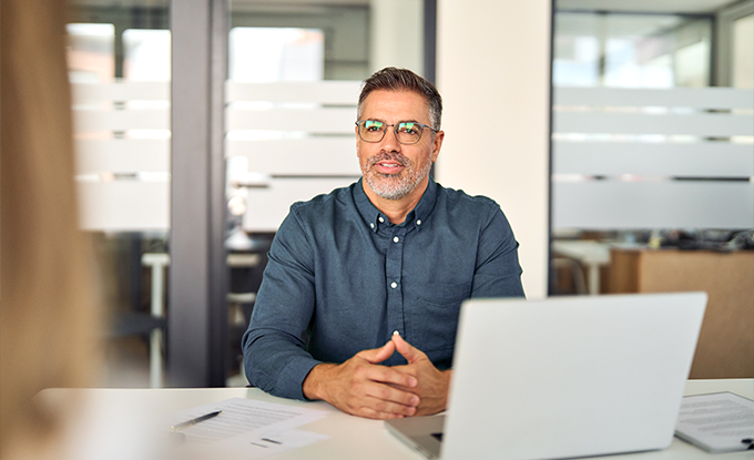 Ein Mann mit Brille und kurzem, grauem Bart sitzt an einem Schreibtisch in einem modernen Büro und spricht. Vor ihm steht ein Laptop, und er wirkt freundlich und professionell.
